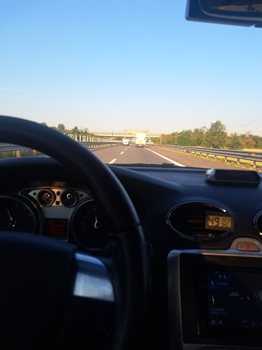 De foto toont het uitzicht vanuit de bestuurdersstoel van een auto op een snelweg. Een deel van het stuur is zichtbaar aan de linkerkant, samen met een deel van het dashboard en de instrumenten. Rechtsonder is een navigatiescherm te zien en een klein digitaal display met de cijfers '49' en '22%'. Voor de auto strekt een meerbaans snelweg zich uit, omzoomd door vangrails en weelderige groene bomen en struiken. Verderop rijden enkele voertuigen, waaronder een lichte vrachtwagen of busje. In de ver