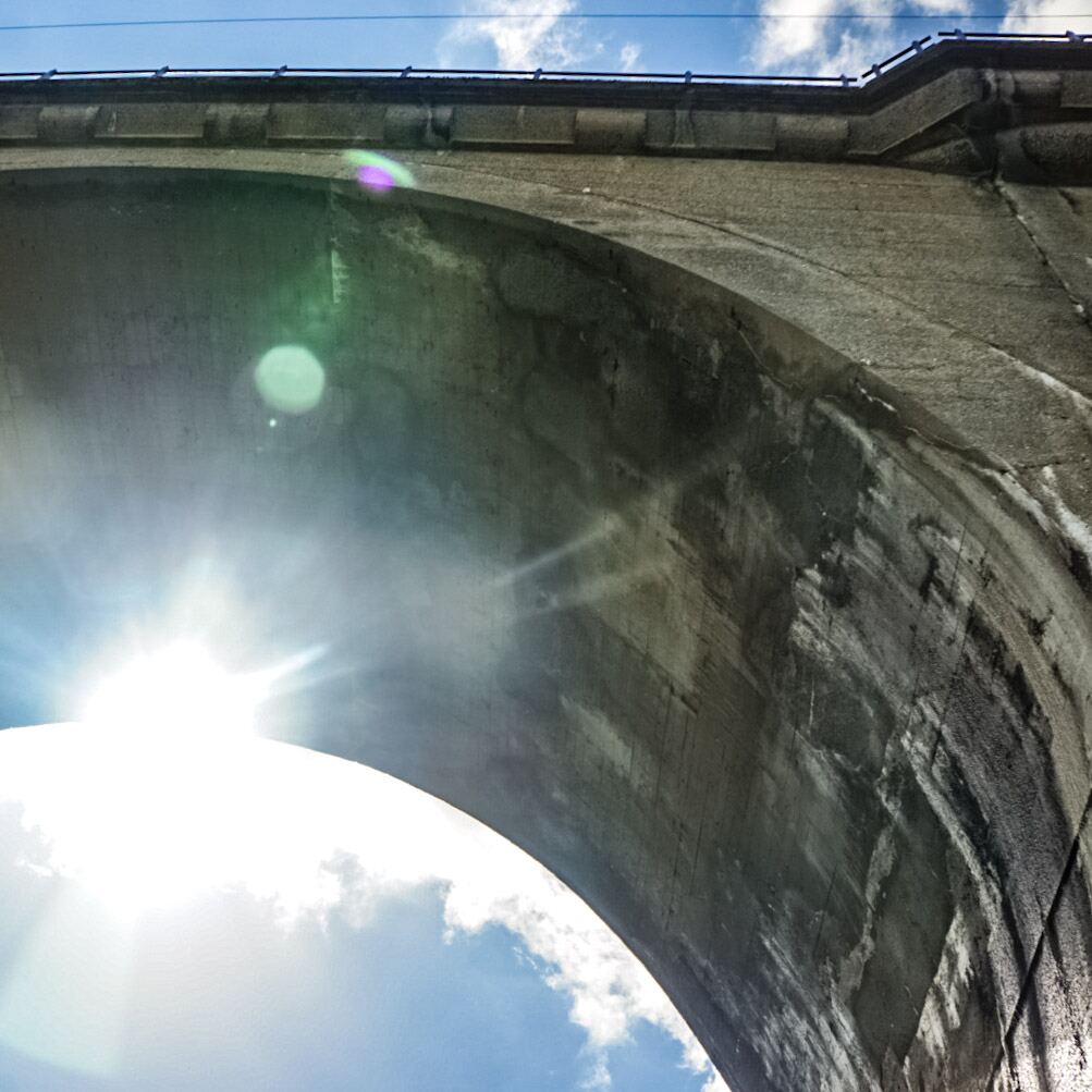 Een foto genomen van onder een betonnen viaduct, met de zon die fel schijnt en lensflares veroorzaakt. De boog van het viaduct domineert de bovenkant van de foto, terwijl de helderblauwe lucht met enkele witte wolken zichtbaar is onder de boog en aan de zijkanten.