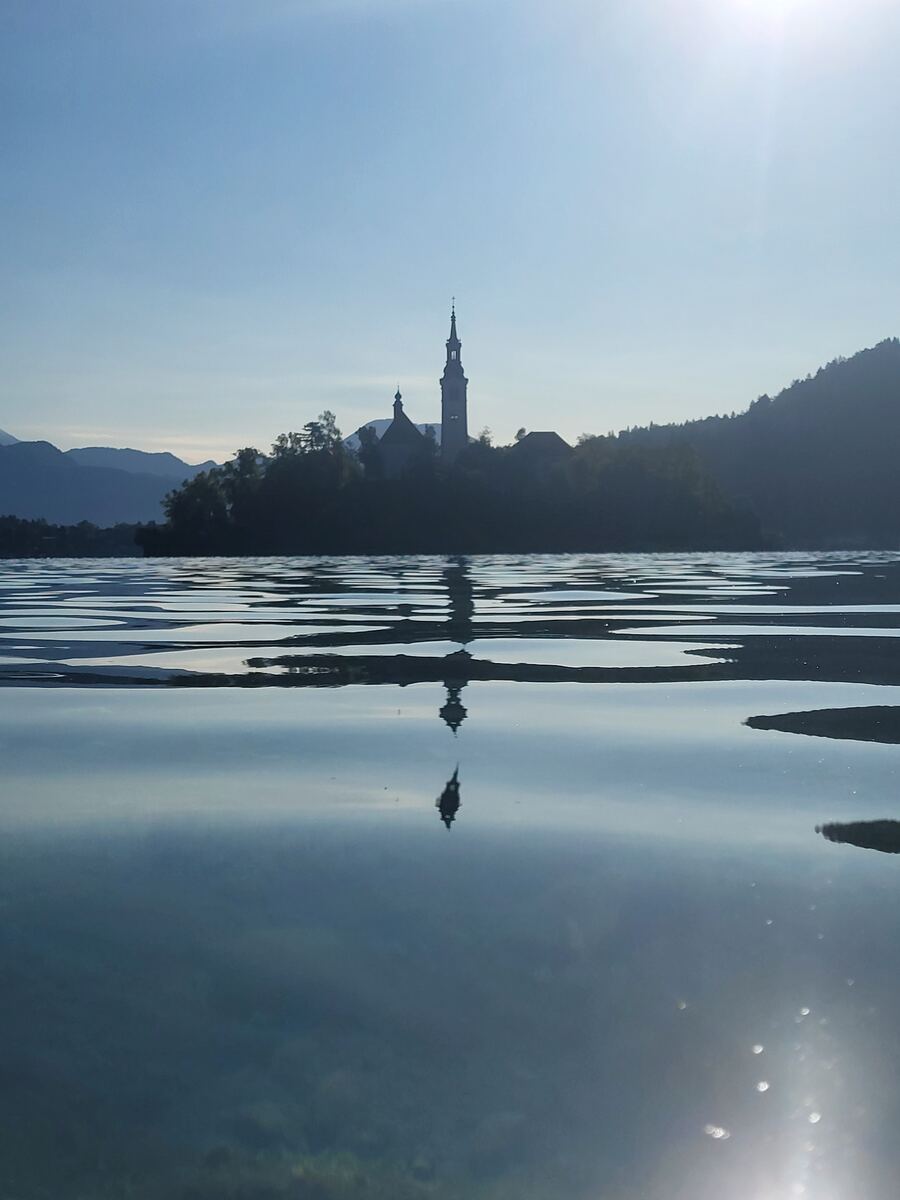 Een silhouet van een kerk met een hoge toren op een eiland in een meer, met een reflectie in het water. Op de achtergrond zijn bergen en beboste heuvels te zien. De zon schijnt fel aan de rechterbovenkant van de foto, wat zorgt voor een lichte gloed op het water.
