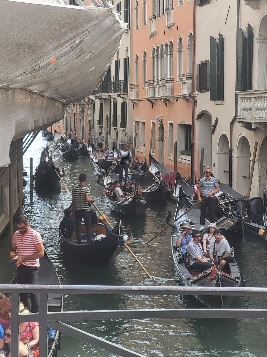 Een smalle gracht in Venetië met meerdere gondels, waarvan sommige met passagiers en gondeliers. De gebouwen langs de gracht hebben traditionele Italiaanse architectuur met ramen, balkons en luiken. Een deel van de gracht is overdekt met een wit zeil of doek.