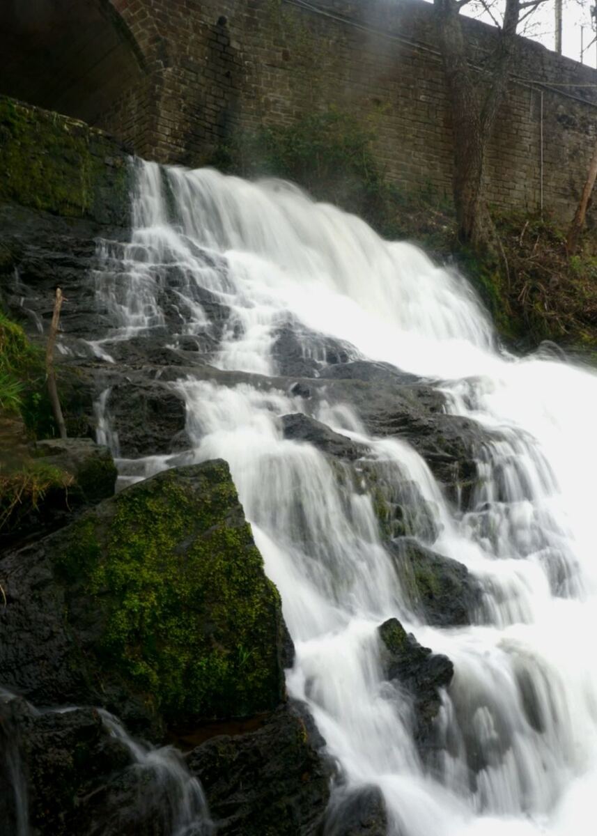 Een waterval stroomt over donkere, met mos begroeide rotsen. Op de achtergrond is een stenen muur met een boog zichtbaar, waarschijnlijk een brug of viaduct.