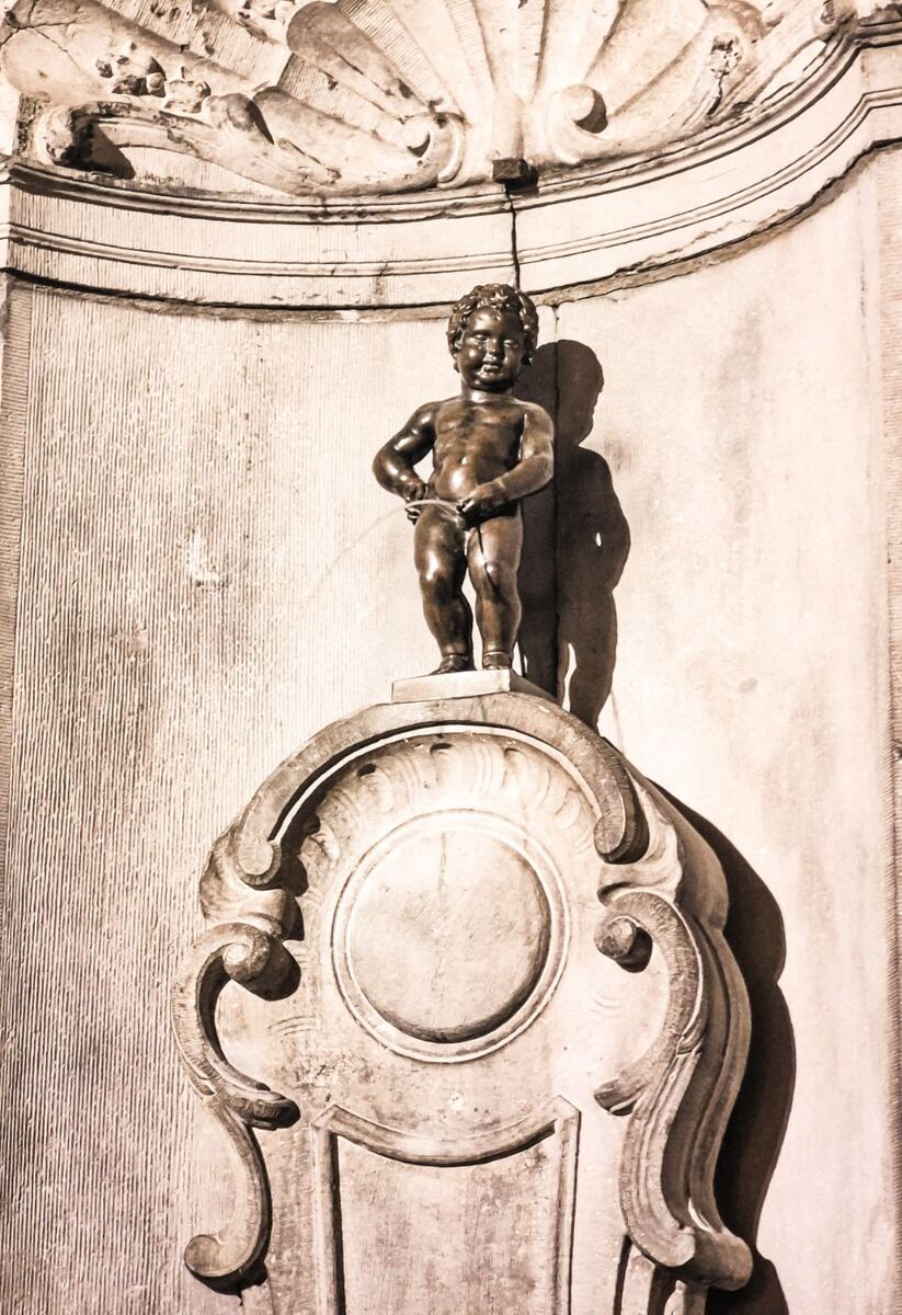 Een sepia-getinte foto van het standbeeld Manneken Pis, een bronzen beeldje van een plassende jongen, staand op een rijk versierde stenen sokkel. Achter het beeld is een muur met een schelpvormig ornament zichtbaar. Er is een duidelijke schaduw van het beeld op de muur.