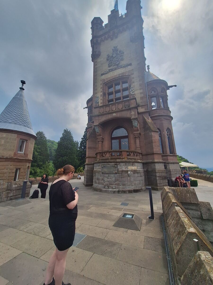 Een foto van een imposant kasteelachtig gebouw met een hoge toren, gezien vanaf een geplaveid terras. Links staat een kleiner gebouw met een kegelvormig dak. Er zijn enkele mensen zichtbaar op het terras, waaronder een vrouw op de voorgrond die op haar telefoon kijkt. De lucht is deels bewolkt en de zon schijnt door de wolken.