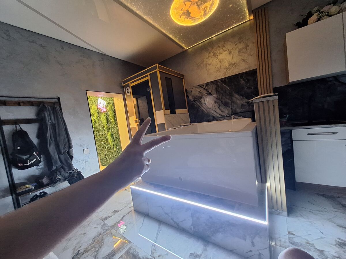 Een moderne badkamer met een groot wit bad, een douchecabine en een opvallend plafond met een afbeelding van een planeet en sterren. Een hand met twee opgestoken vingers is prominent in beeld. De muren zijn deels van marmer en deels van een grijze structuur. Er is ook een deur met een groene, mosachtige bekleding en een neonreclame.