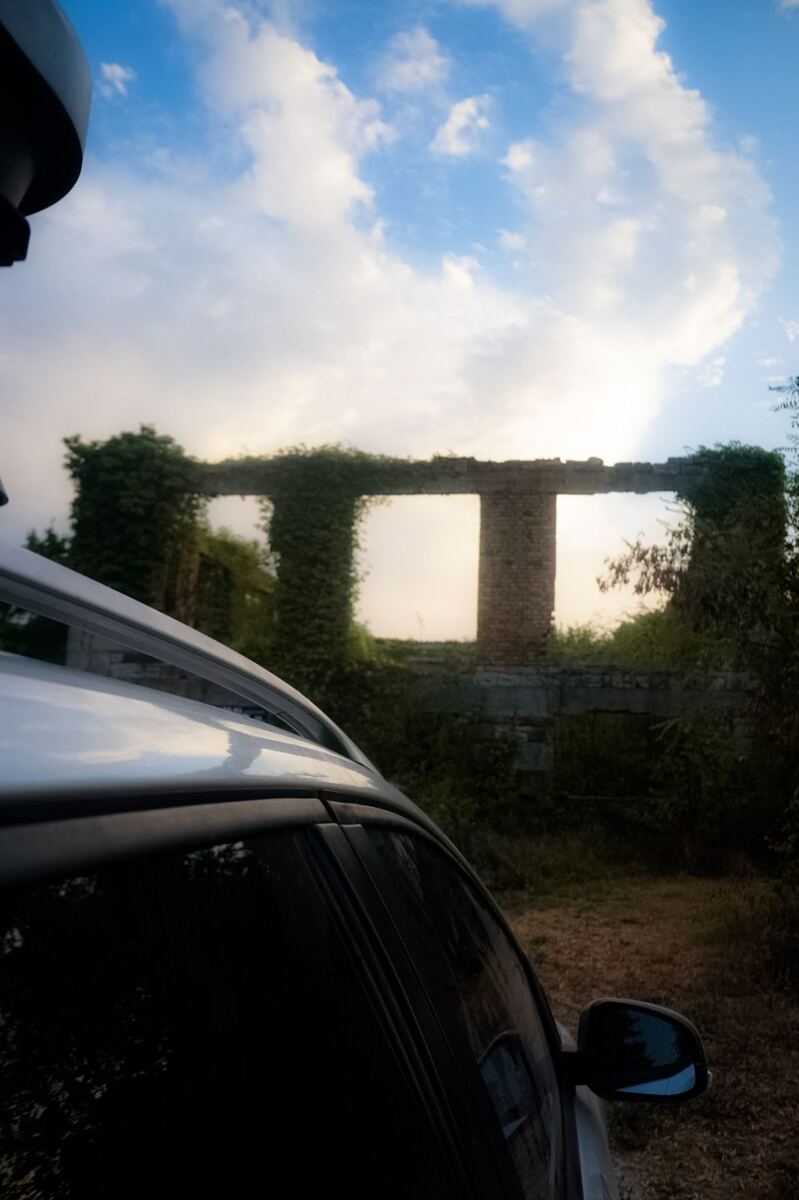 De foto toont de zijkant van een witte auto met dakrails, met op de achtergrond de overblijfselen van een stenen gebouw, deels overwoekerd met klimop. De lucht erboven is blauw met witte wolken, en de zon schijnt door de open vensters van de ruïne.