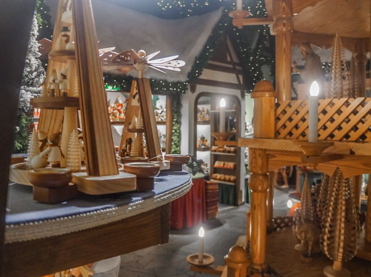 Een interieurfoto van een winkel met houten kerstdecoraties, waaronder piramides met figuren en kaarsen. Op de achtergrond zijn meer kerstartikelen en een gevel met kerstverlichting te zien.