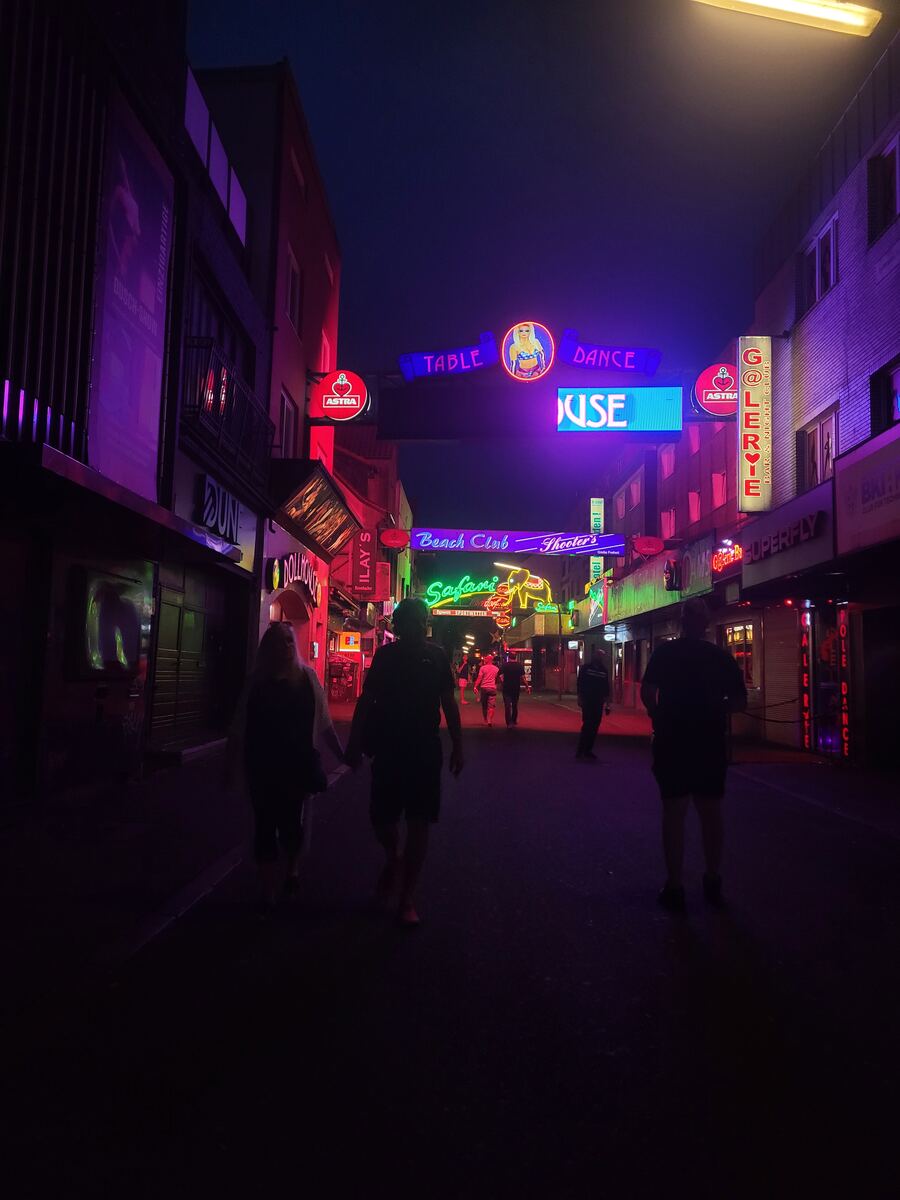 Een donkere straat verlicht door diverse neonreclames in roze, paarse, groene en blauwe tinten. Verschillende mensen lopen over de straat, waarvan twee personen op de voorgrond hand in hand lopen. Aan weerszijden van de straat staan gebouwen met ramen en reclameborden, waaronder 'Astra' en 'Galerie'.