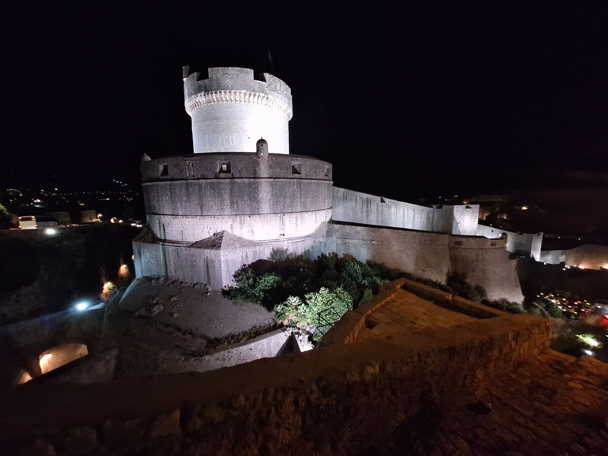 Een nachtfoto van een verlichte ronde toren en een deel van de stadsmuren van Dubrovnik, met op de achtergrond de lichtjes van de stad.