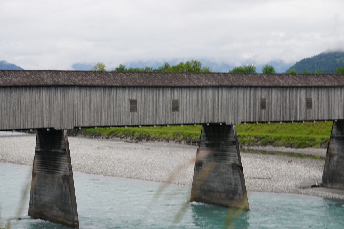 Een houten overdekte brug met een pannendak over een rivier met turquoise water en een kiezelstrand. Op de achtergrond zijn groene bomen en bergen te zien onder een bewolkte lucht.