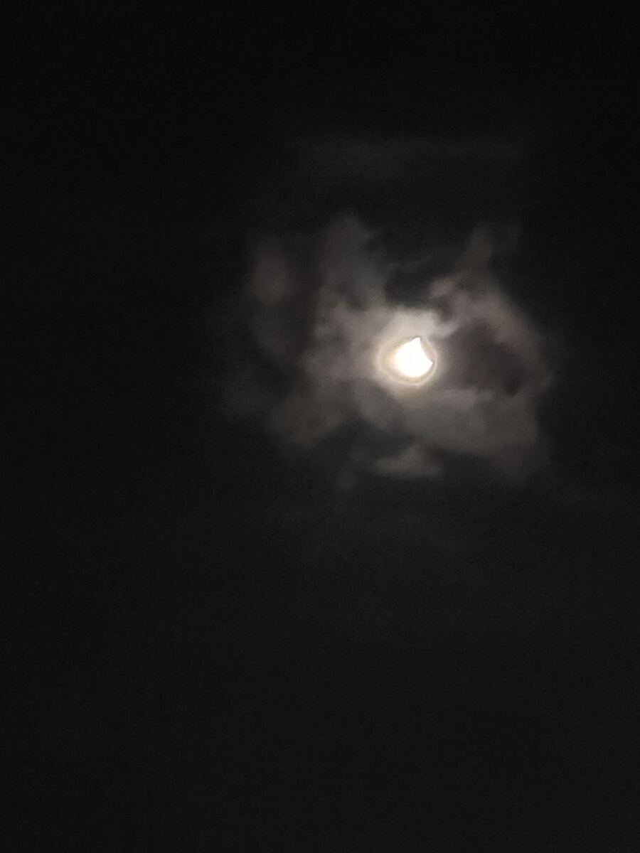 Een donkere nachtelijke hemel met de maan die deels zichtbaar is door een laag wolken. De maan is helder en heeft een sikkelvormige uitsnede, wat duidt op een maansverduistering of een gedeeltelijke bedekking door een ander hemellichaam. De wolken rondom de maan zijn diffuus en lichtgrijs, terwijl de rest van de lucht pikzwart is.