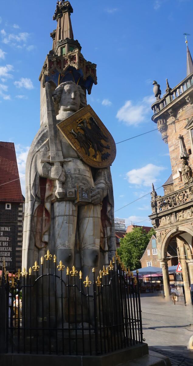 Een standbeeld van Roland, een ridderfiguur, staat prominent in het midden van de foto onder een blauwe lucht met enkele wolken. Het standbeeld is van steen en draagt een schild met een tweekoppige adelaar. Rondom het standbeeld staat een zwart hek met gouden lelies. Rechts van het standbeeld is een deel van een historisch gebouw met bogen en versieringen zichtbaar. Op de achtergrond zijn ook andere gebouwen met rode daken en wat groen te zien.