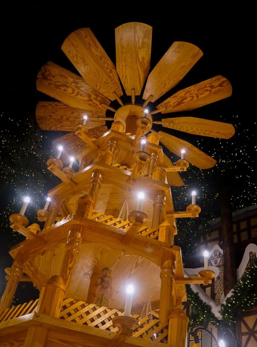 Een grote, houten kerstpiramide met meerdere etages en draaiende wieken, verlicht met elektrische kaarsen. Op de achtergrond zijn donkere gebouwen te zien met kerstverlichting en een donkere lucht met kleine lichtjes die op sterren lijken.
