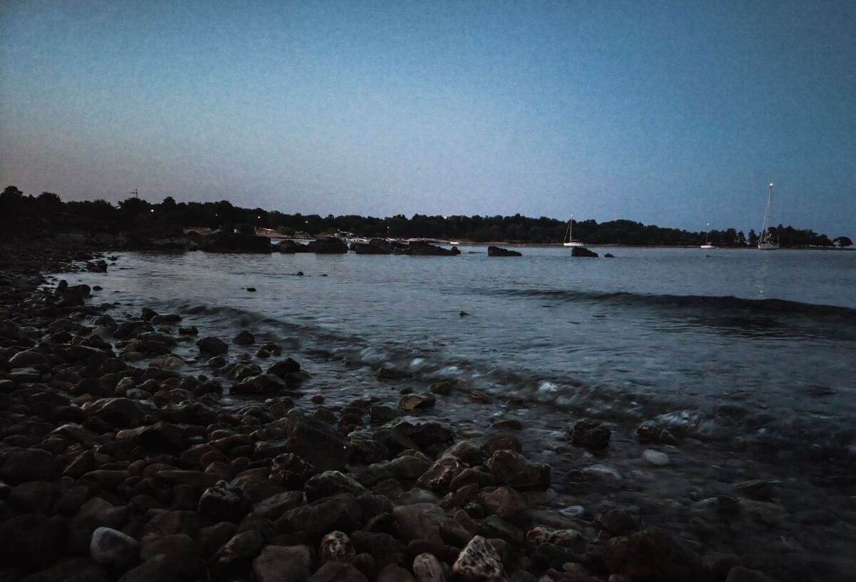 Een kustlijn bij zonsopkomst of schemering, met een rotsachtig strand op de voorgrond en kalm water dat de kustlijn raakt. Aan de horizon zijn bomen en enkele zeilboten zichtbaar onder een heldere, blauwgrijze lucht.