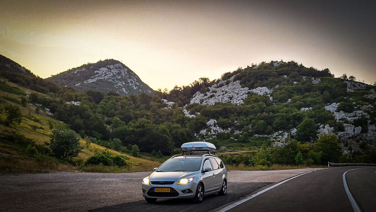 Een zilverkleurige Ford Focus stationwagen met een dakkoffer staat geparkeerd langs een bochtige weg in een bergachtig landschap. De zon staat laag en werpt een zacht, geel licht over de heuvels die begroeid zijn met bomen en rotsformaties. De lucht is deels bewolkt en de weg heeft witte belijning.