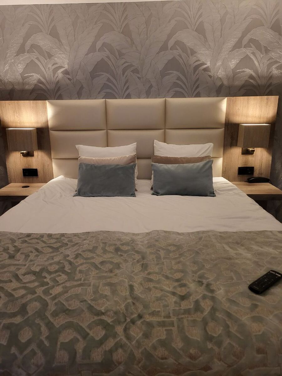 Een hotelkamer met een opgemaakt tweepersoonsbed, twee nachtkastjes met lampen en een telefoon. De muur achter het bed heeft behang met een bladmotief.
