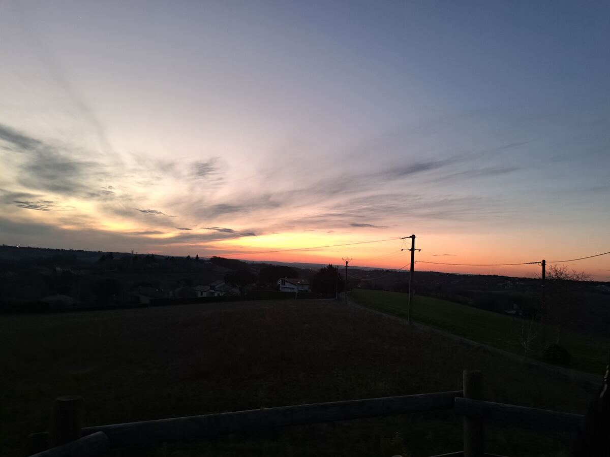 Een weids uitzicht over een glooiend landschap bij zonsondergang, met de lucht die van geel en oranje naar paars en blauw kleurt. Op de voorgrond zijn donkere velden te zien, met enkele huizen en bomen in de verte. Elektriciteitspalen en draden lopen door het midden van de foto, en een houten hekwerk is zichtbaar aan de onderrand.