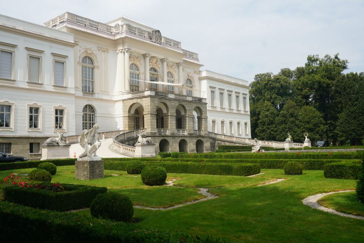 Een wit barok gebouw met een grote tuin ervoor. De tuin heeft strak gesnoeide hagen en beelden. Het gebouw heeft een centrale ingang met bogen en een trap die naar boven leidt.