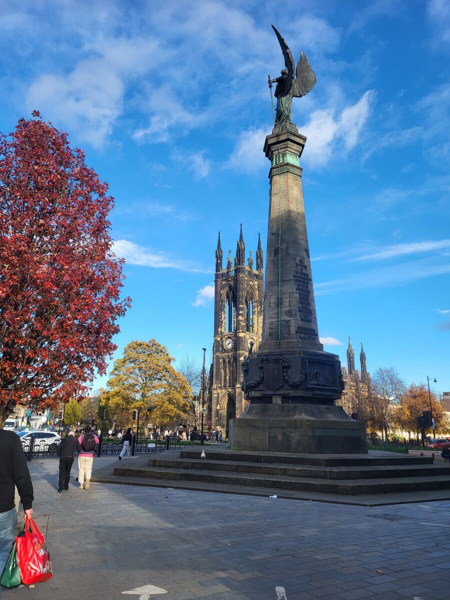 Een stenen monument met een gevleugeld beeld op een sokkel staat centraal, met daarachter een grote neogotische kerk met een klok. Links en in de verte staan bomen met herfstkleuren. Op de voorgrond is een geplaveid plein te zien waar mensen lopen en een meeuw op de trap van het monument zit. De lucht is blauw met witte wolken.