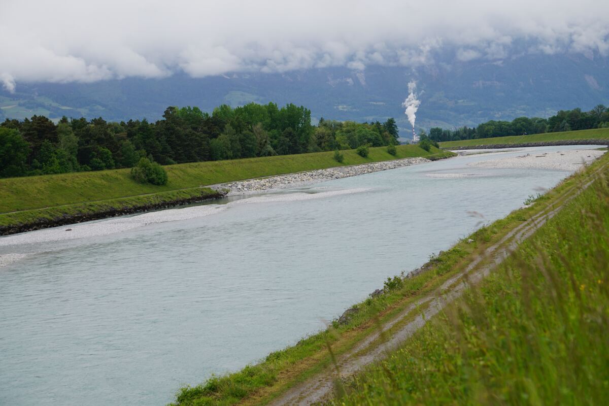 Een brede rivier met lichtblauw water stroomt door een vallei. Aan beide zijden van de rivier zijn groene oevers met gras en bomen. Op de achtergrond zijn bergen te zien die deels in de wolken liggen. Rechts in de verte is een schoorsteen met rookpluim zichtbaar.