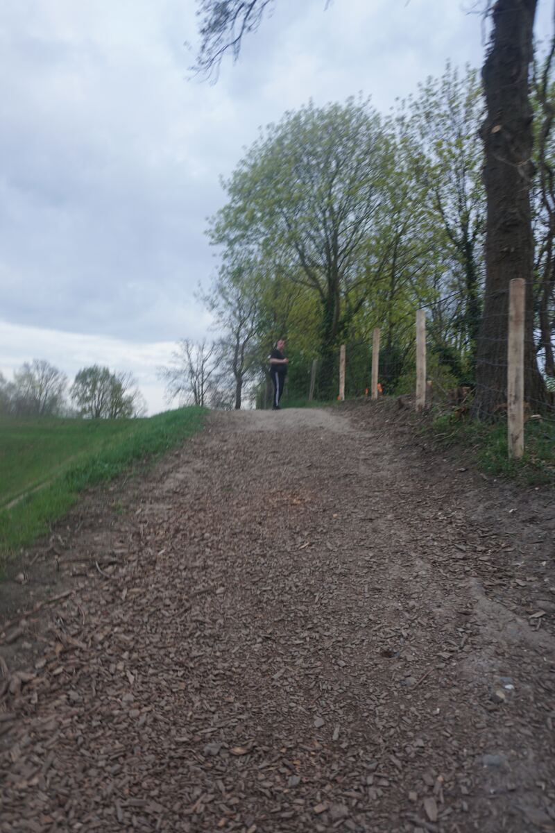 Een persoon staat op een onverhard pad van houtsnippers, omringd door bomen en groen. De lucht is bewolkt en grijs. Aan de rechterkant van het pad staan houten palen met gaas.
