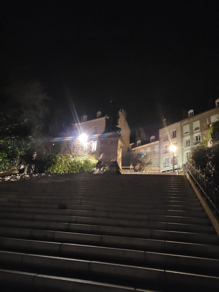 Een lange, brede stenen trap leidt omhoog naar een verlichte straat met gebouwen en bomen. Links van de trap staat een monument of standbeeld. De scène is 's nachts opgenomen, met straatverlichting die de omgeving verlicht.