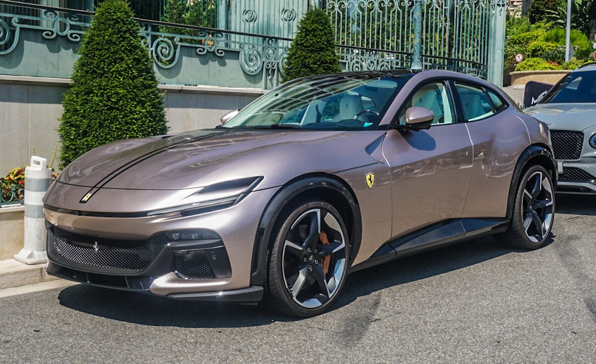 Een beige-grijze Ferrari Purosangue staat geparkeerd op een geasfalteerde straat. De auto heeft zwarte velgen en oranje remklauwen. Achter de Ferrari is een deel van een andere luxe auto, waarschijnlijk een Bentley, zichtbaar. Op de achtergrond is een sierlijk metalen hek met groen struikgewas te zien. De zon schijnt helder en er zijn duidelijke schaduwen op de weg.