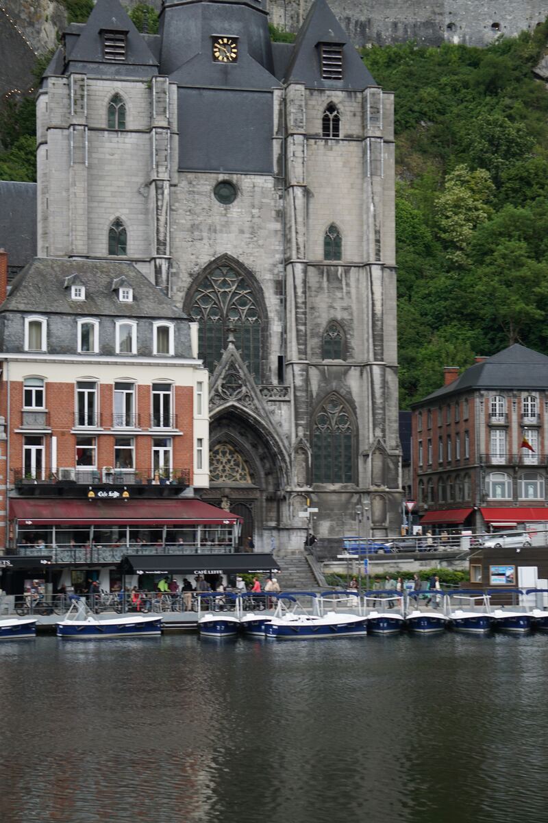 Een verticale foto toont de Collegiale kerk van Dinant met zijn kenmerkende uivormige koepel en klok, gelegen aan de Maas. Op de voorgrond liggen meerdere kleine, blauw-witte plezierbootjes aangemeerd. Aan de oever staan gebouwen met rode daken en bakstenen gevels, waaronder een restaurant met een rood zonnescherm en de naam 'Café Leffe'. Achter de kerk is een groene, beboste heuvel zichtbaar.