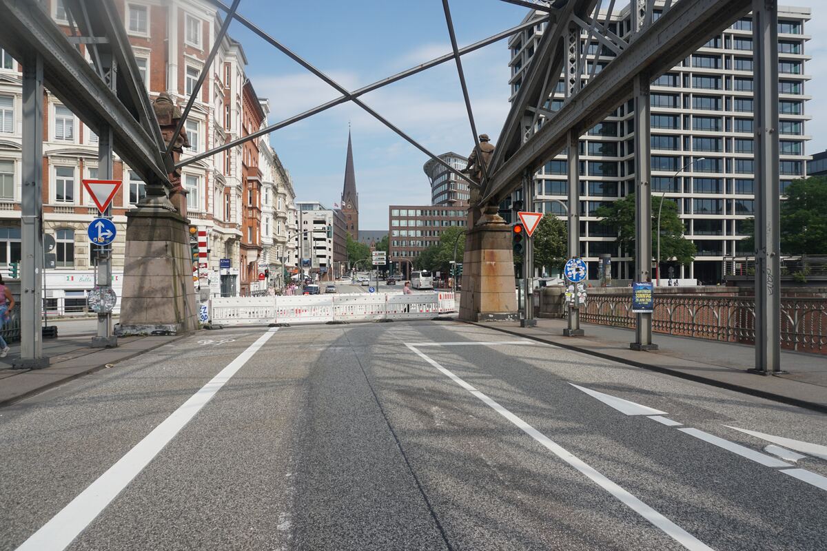 Een brede stadsstraat gezien onder een metalen brugconstructie. Links en rechts van de weg staan diverse gebouwen, waaronder oudere bakstenen panden en moderne kantoorgebouwen. In de verte is een spitse kerktoren zichtbaar. Op de weg zijn witte rijstrookmarkeringen en tijdelijke witte bouwbarrières geplaatst. Verkeersborden en een rood verkeerslicht zijn ook te zien.