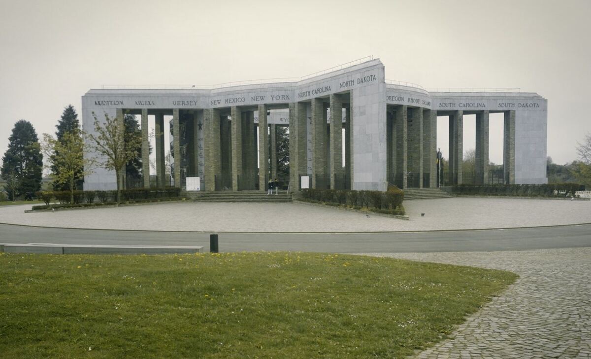 Een breed overzicht van het Mémorial du Mardasson, een stervormig monument met zuilen en namen van Amerikaanse staten op de bovenrand. De lucht is bewolkt en de omgeving bestaat uit grasvelden en bomen.