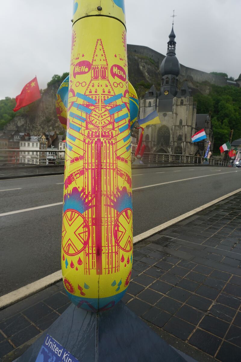 Een close-up van een felgele, beschilderde paal met roze en blauwe patronen, waaronder een gestileerde kerk en de tekst 'Hello'. Op de achtergrond is een brug te zien met vlaggen van verschillende landen, een kerk met een donkere koepel en een rotswand met begroeiing.