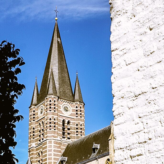 Een foto van een kerk met een hoge, spitse toren en twee kleinere torens, gemaakt vanuit een laag standpunt. De toren is opgebouwd uit afwisselende lagen van baksteen en lichtere stenen, met klokken en ramen. Rechts is een deel van een witte, ruwe muur zichtbaar. De lucht is helderblauw met enkele lichte wolken. Links is een donker silhouet van bladeren te zien.