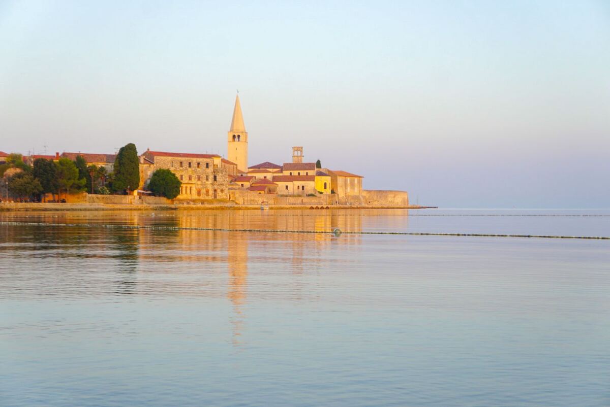 Een avondopname van de kustlijn van Poreč, Kroatië, met gebouwen en een kerktoren die reflecteren in het kalme water. De lucht is helder met een zachte overgang van lichtblauw naar roze. De zon staat laag, waardoor de gebouwen een warme, gouden gloed hebben.