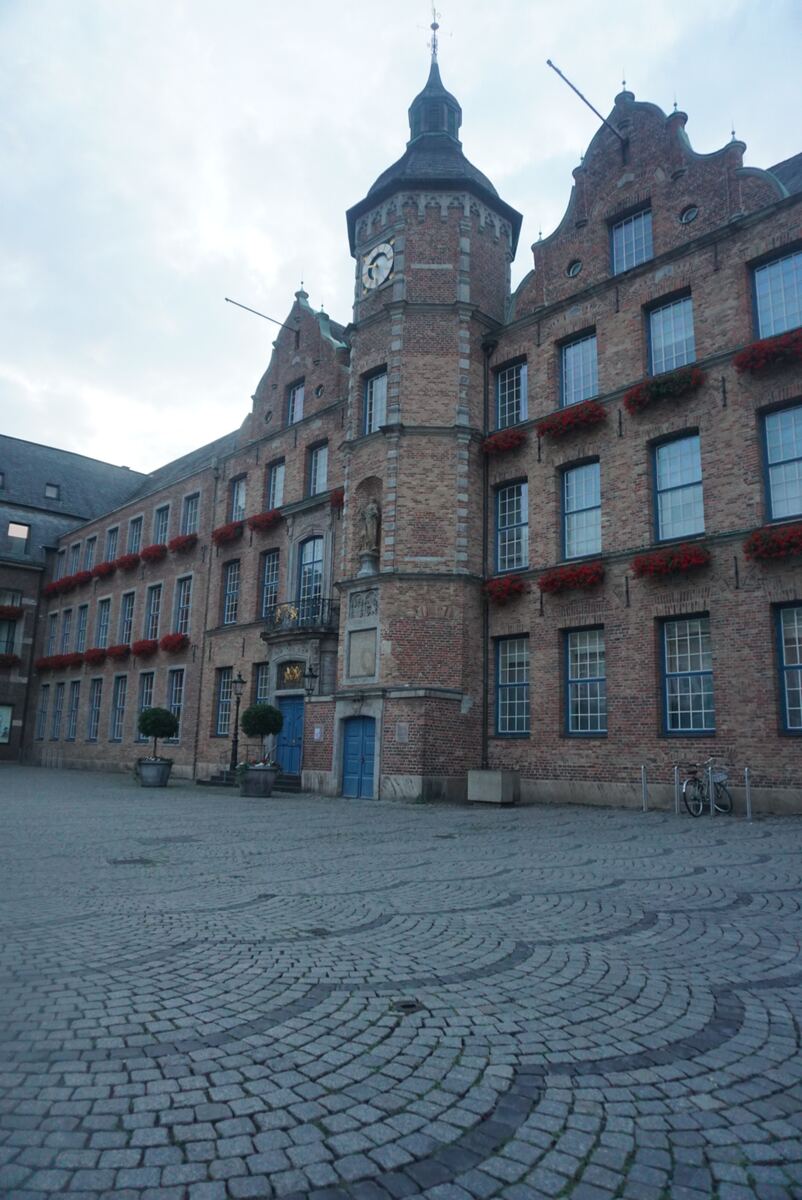 Een groot, historisch bakstenen gebouw met een klokkentoren en meerdere verdiepingen, versierd met rode bloembakken. De gevel heeft veel ramen met blauwe kozijnen en er zijn twee blauwe deuren zichtbaar. De voorgrond bestaat uit een geplaveid plein met een patroon van kasseien. Aan de rechterkant staan twee fietsen geparkeerd.