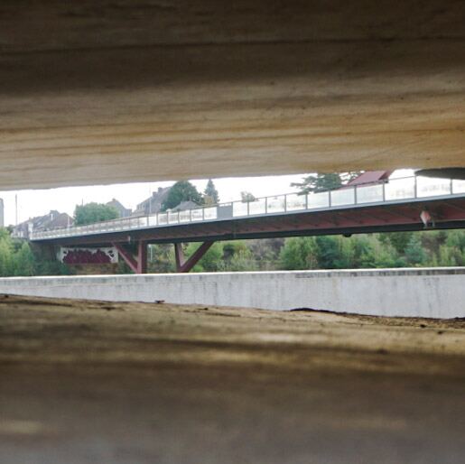 Een foto genomen onder een brug door, met een andere brug op de achtergrond die over water lijkt te lopen. De achtergrond toont huizen en bomen.