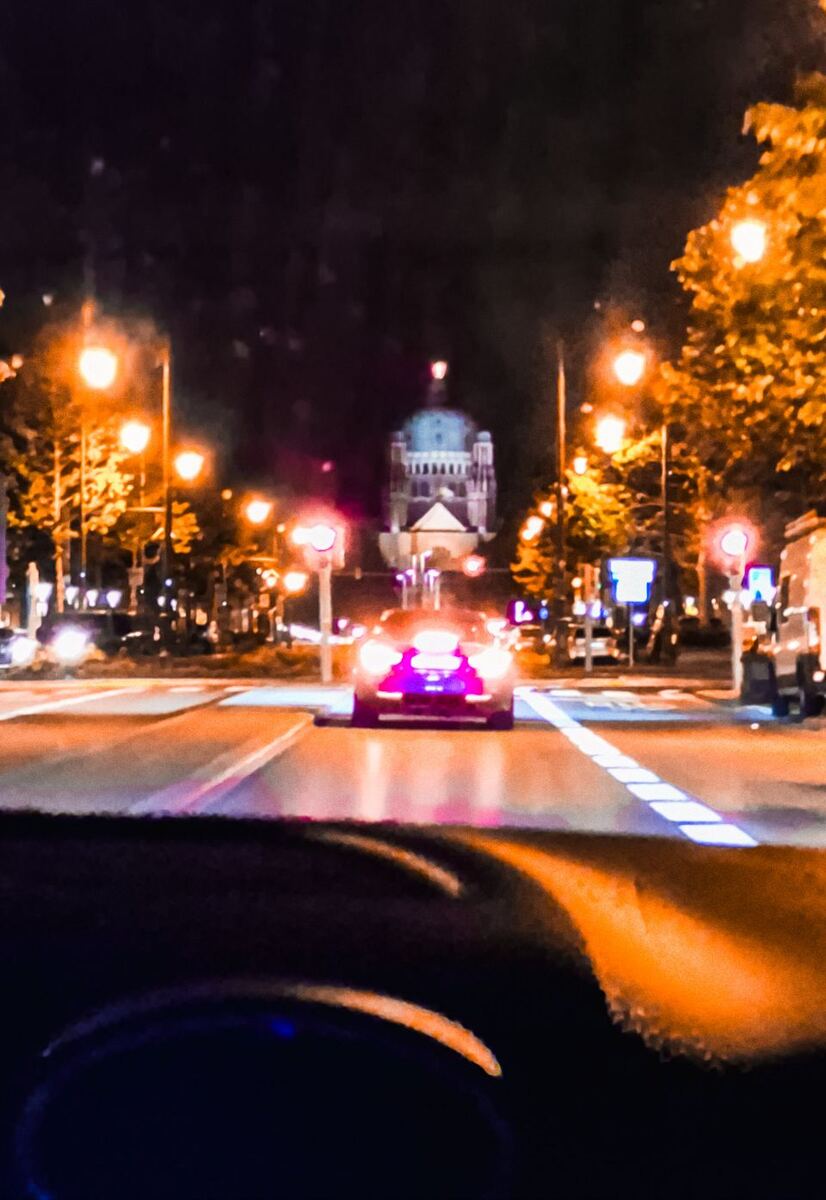 Een nachtelijke opname vanuit een auto, gericht op een straat met een gebouw met een koepel op de achtergrond. Er zijn straatverlichting en verkeerslichten zichtbaar, evenals een auto met felle koplampen die voor ons rijdt. De voorruit van onze auto is deels zichtbaar aan de onderkant van de foto.
