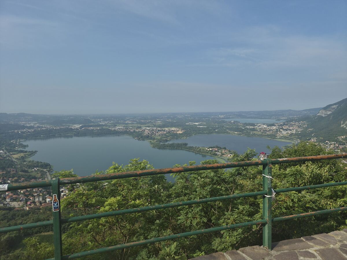 Een panoramisch uitzicht over een uitgestrekt landschap met twee meren, omringd door bebouwing en groene heuvels en bergen, gezien vanaf een hoog uitkijkpunt. Op de voorgrond is een groen, deels roestig metalen hekwerk te zien, met daarachter weelderig groen gebladerte. Aan het hek hangt links een sticker van een gevleugelde bierfles en rechts zijn enkele kleine witte lintjes of labels bevestigd. De lucht is lichtblauw en helder, met een lichte waas aan de horizon.