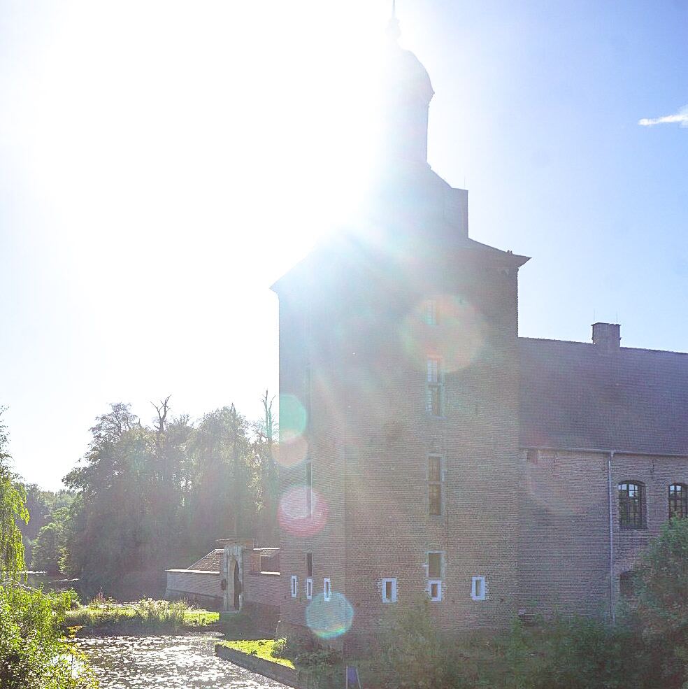 Een bakstenen kasteel met een toren en een koepeldak, deels verlicht door een felle zon die lensflares veroorzaakt. Op de voorgrond is een waterloop met reflecties van het zonlicht te zien, omringd door groen. De lucht is blauw en helder.
