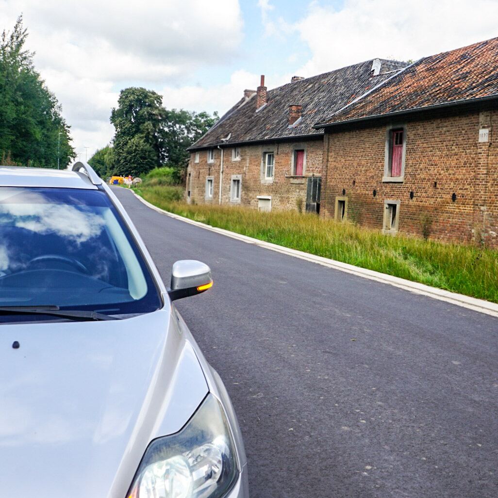 Een zilverkleurige auto staat geparkeerd langs een smalle asfaltweg. Aan de rechterkant van de weg staan twee oude, bakstenen boerderijen met rode dakpannen en dichte rode luiken. De huizen hebben kleine ramen en er groeit hoog gras langs de gevels. Op de achtergrond zijn bomen en een bewolkte lucht te zien.
