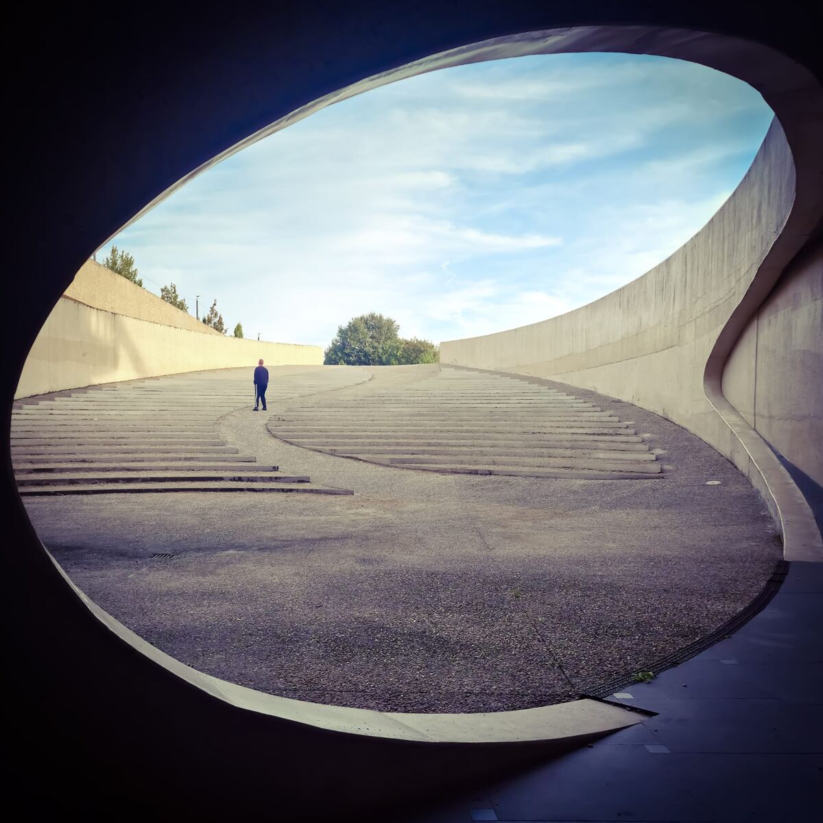 Een man loopt over een pad in een openluchttheater of amfitheater, omringd door betonnen muren en rijen zitplaatsen. De scène wordt gezien door een ovale opening.