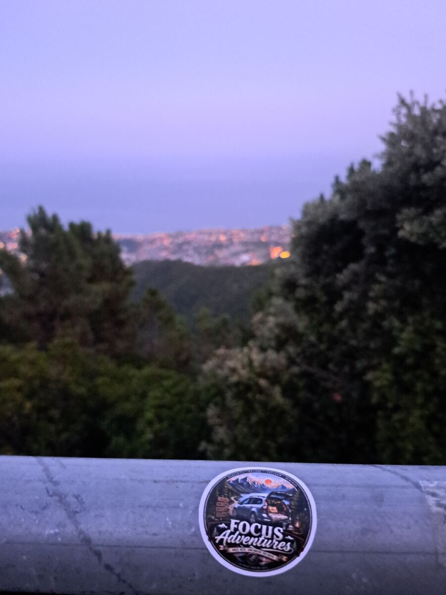 De foto toont een avondlandschap vanaf een hoge positie. Op de voorgrond is een grijze metalen reling zichtbaar met daarop een ronde sticker met de tekst 'Focus Adventures' en een afbeelding van twee auto's in een bergachtige, bosrijke omgeving bij zonsopgang of zonsondergang. De reling en sticker zijn scherp. Daarachter zijn wazige groene bomen en struiken te zien. Op de achtergrond strekt zich een onscherpe stad uit met talloze brandende lichtjes, die overgaan in een donkere watermassa (waarsc