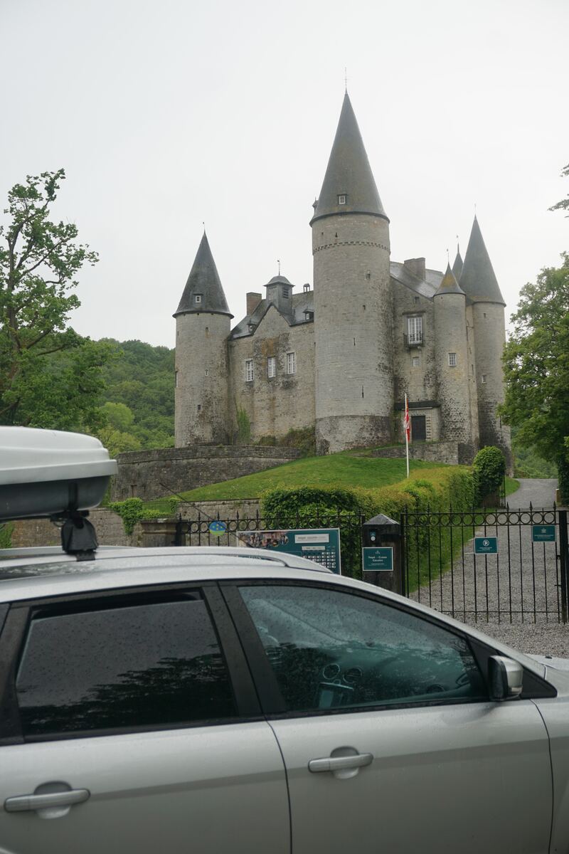Een zilverkleurige auto met een dakkoffer staat op de voorgrond, met op de achtergrond een groot stenen kasteel met meerdere torens en puntige daken. Het kasteel is omgeven door groen en een heuvelachtig landschap onder een bewolkte lucht. Voor het kasteel is een zwart hek zichtbaar met informatieborden.