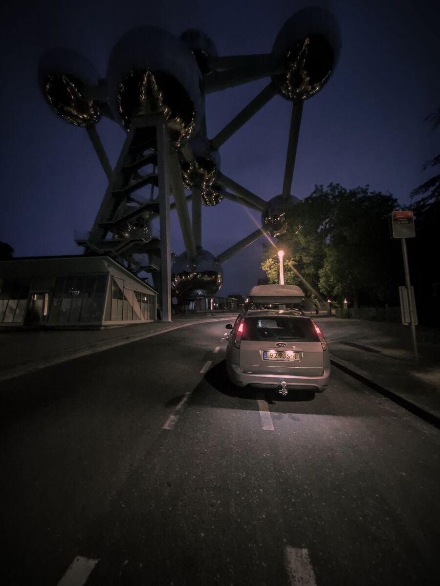 Een Ford Focus stationwagen staat geparkeerd op een weg voor het Atomium in Brussel, gefotografeerd in de schemering of vroege ochtend. De lucht is donkerblauw en de verlichting van het Atomium en een straatlantaarn zijn zichtbaar. De auto heeft een dakkoffer en een trekhaak.