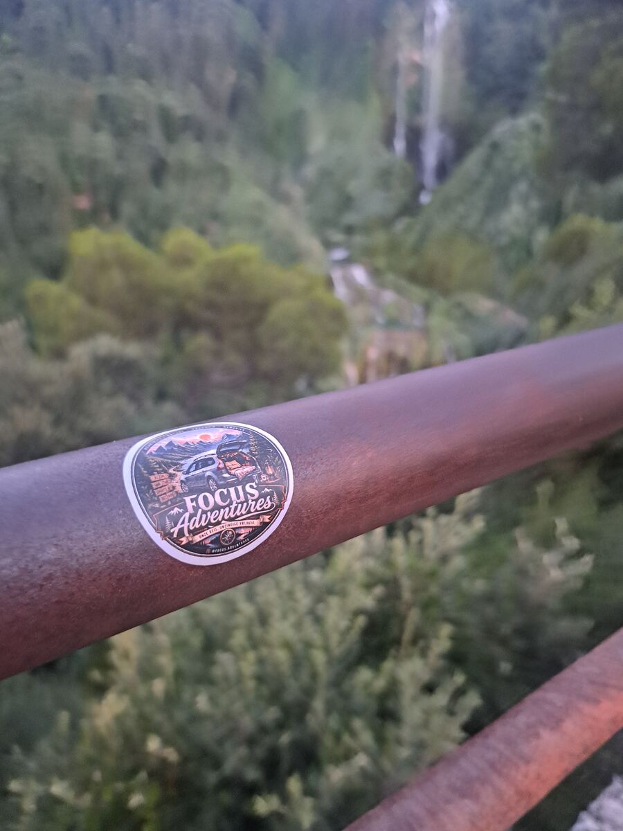 Een close-up van een roestbruine metalen railing met daarop een ronde, kleurrijke sticker. De sticker toont een illustratie van een auto met open kofferbak in een berglandschap bij zonsondergang, met de tekst 'FOCUS Adventures' en 'ONZE REIS, ONZE NIEUWE TRADITIE'. Op de achtergrond is, onscherp, een diepe groene vallei te zien met dichte begroeiing van bomen en struiken. In de verte is een heldere waterval zichtbaar die naar beneden stort, en een kronkelende rivier of beek op de bodem van de va