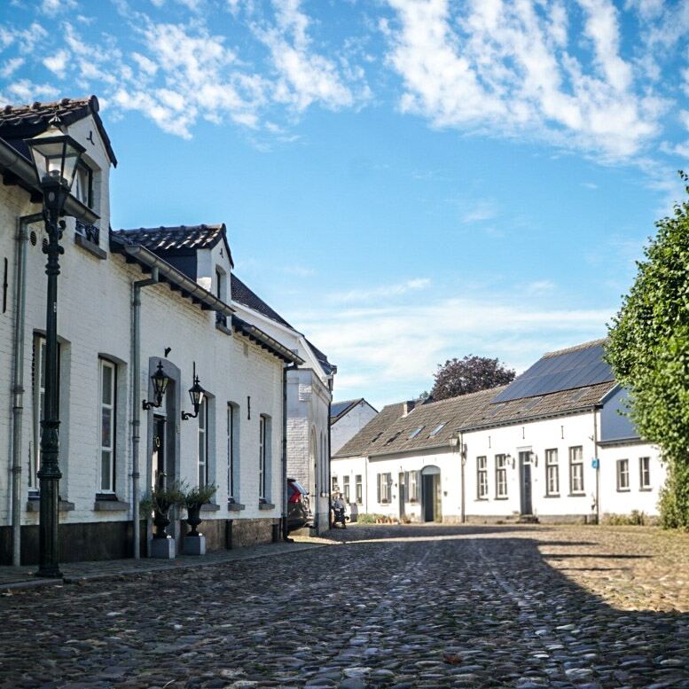 Een geplaveide straat met aan weerszijden witte huizen onder een blauwe lucht met wolkjes. Links staat een klassieke lantaarnpaal en op een van de daken zijn zonnepanelen te zien. De zon werpt schaduwen op de straat.