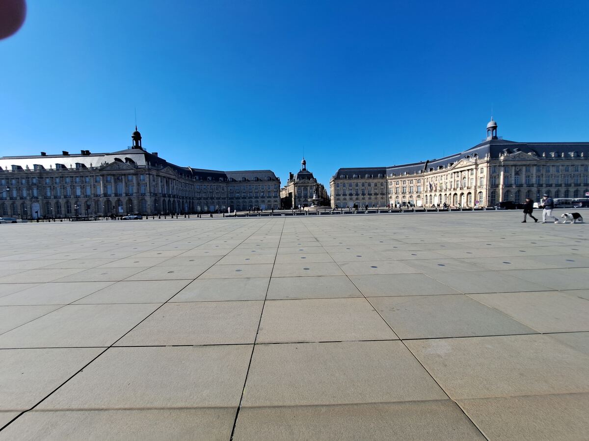 Een weidse foto van een groot, leeg stadsplein met op de achtergrond twee symmetrische, klassieke gebouwen met donkere daken en een kleiner gebouw met een koepel in het midden. De lucht is helderblauw en er zijn enkele mensen met een hond zichtbaar aan de rechterkant van het plein.