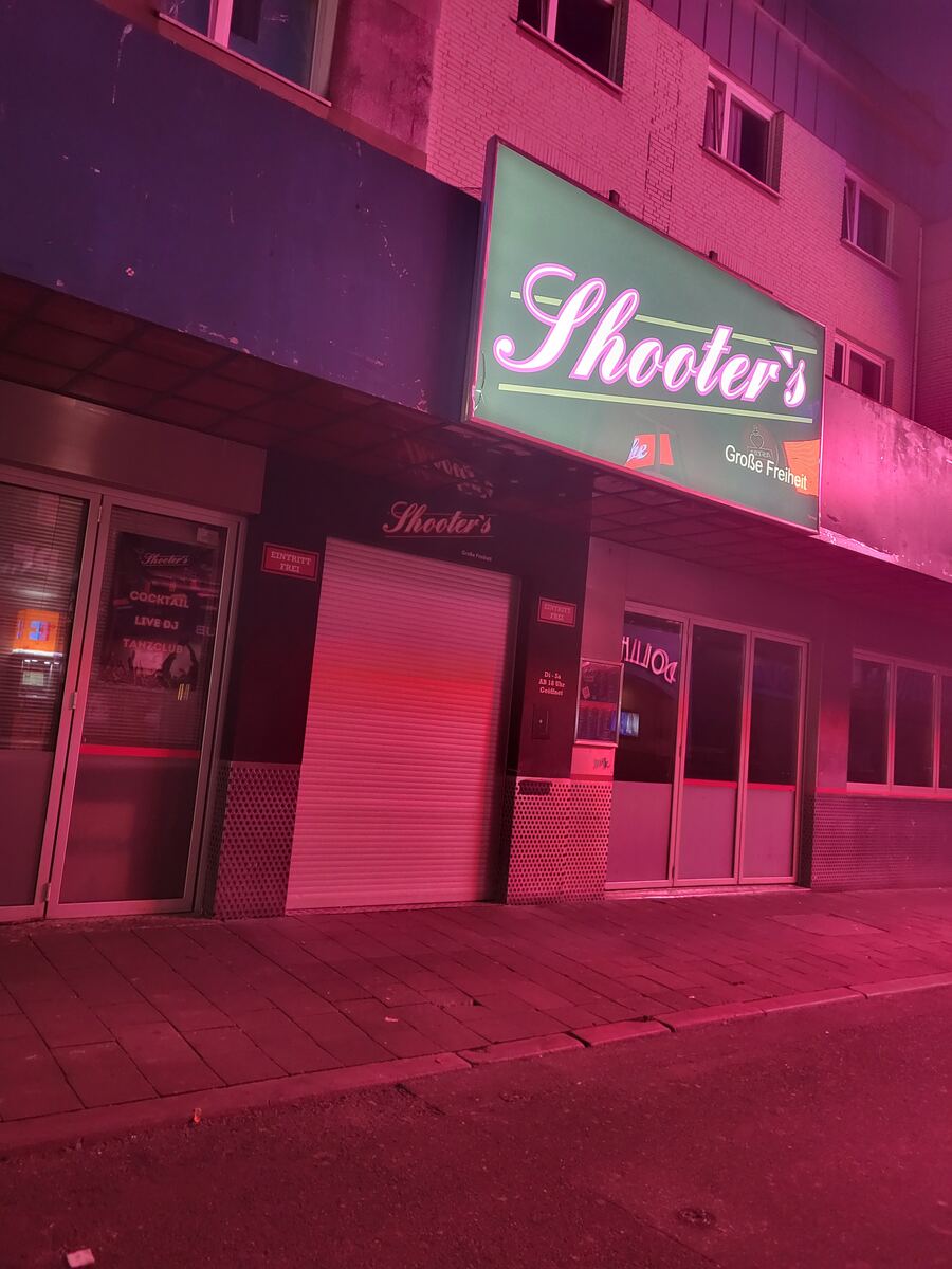Een gevel van een gebouw met een verlicht neonreclamebord van 'Shooter's' in een roze gloed. De straat en de gevel zijn sterk gekleurd door roze licht. Er zijn twee deuren zichtbaar, waarvan één met een gesloten rolluik. Op de ramen zijn teksten te lezen zoals 'Cocktail', 'Live DJ' en 'Tanzclub'. Ook is 'Eintritt Frei' en 'Große Freiheit' te zien.