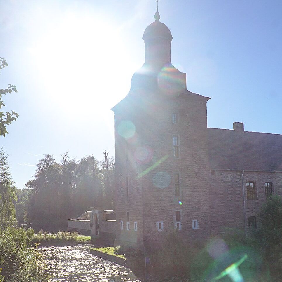 Een foto van een kasteel met een opvallende toren en een slotgracht. De zon schijnt fel en veroorzaakt lensflares. Rondom het kasteel is veel groen te zien.