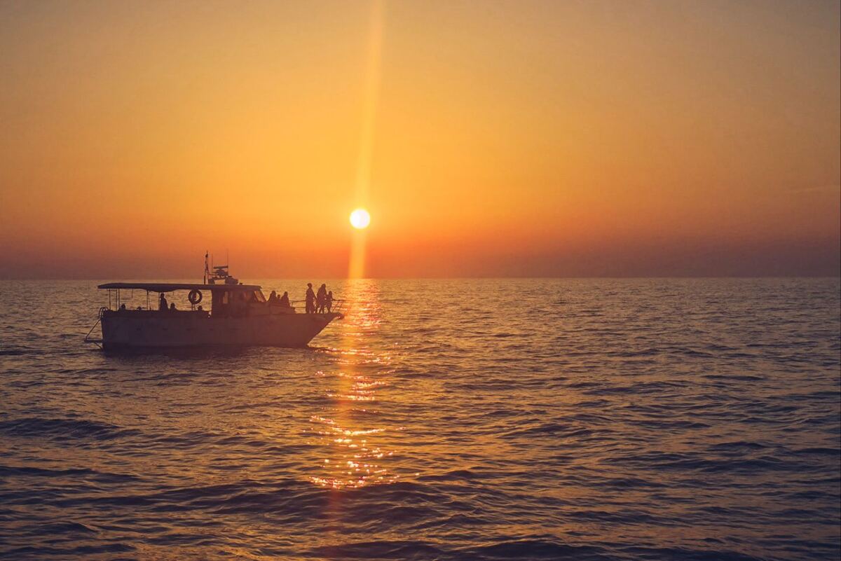 Een boot met mensen aan boord vaart over rustig water tijdens zonsondergang. De zon staat laag aan de horizon en werpt een heldere lichtstraal over het wateroppervlak. De lucht is oranje en geel gekleurd. De boot is in silhouet zichtbaar en er zijn meerdere mensen aan dek te zien.