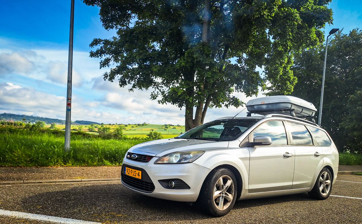 Een witte Ford Focus stationwagen met een dakkoffer staat geparkeerd op een parkeerplaats langs een weg. Op de achtergrond is een groen heuvellandschap te zien onder een deels bewolkte blauwe lucht. Rechts van de auto staat een grote boom en links een lantaarnpaal. De nummerplaat van de auto is Nederlands.