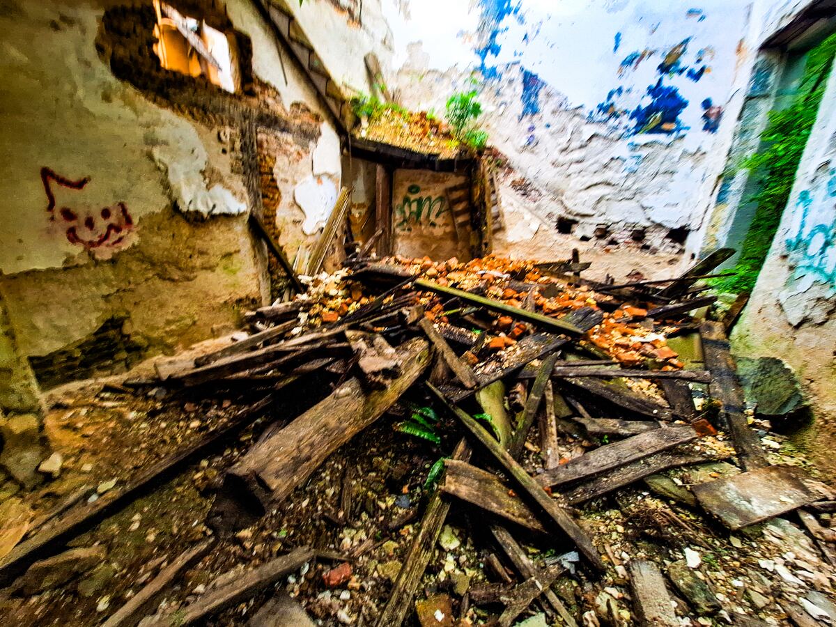 Een interieur van een vervallen gebouw met ingestorte muren en een vloer bedekt met puin, bestaande uit houten planken, bakstenen en aarde. Er is graffiti zichtbaar op de muren en door een opening in de muur is een stukje blauwe lucht te zien.